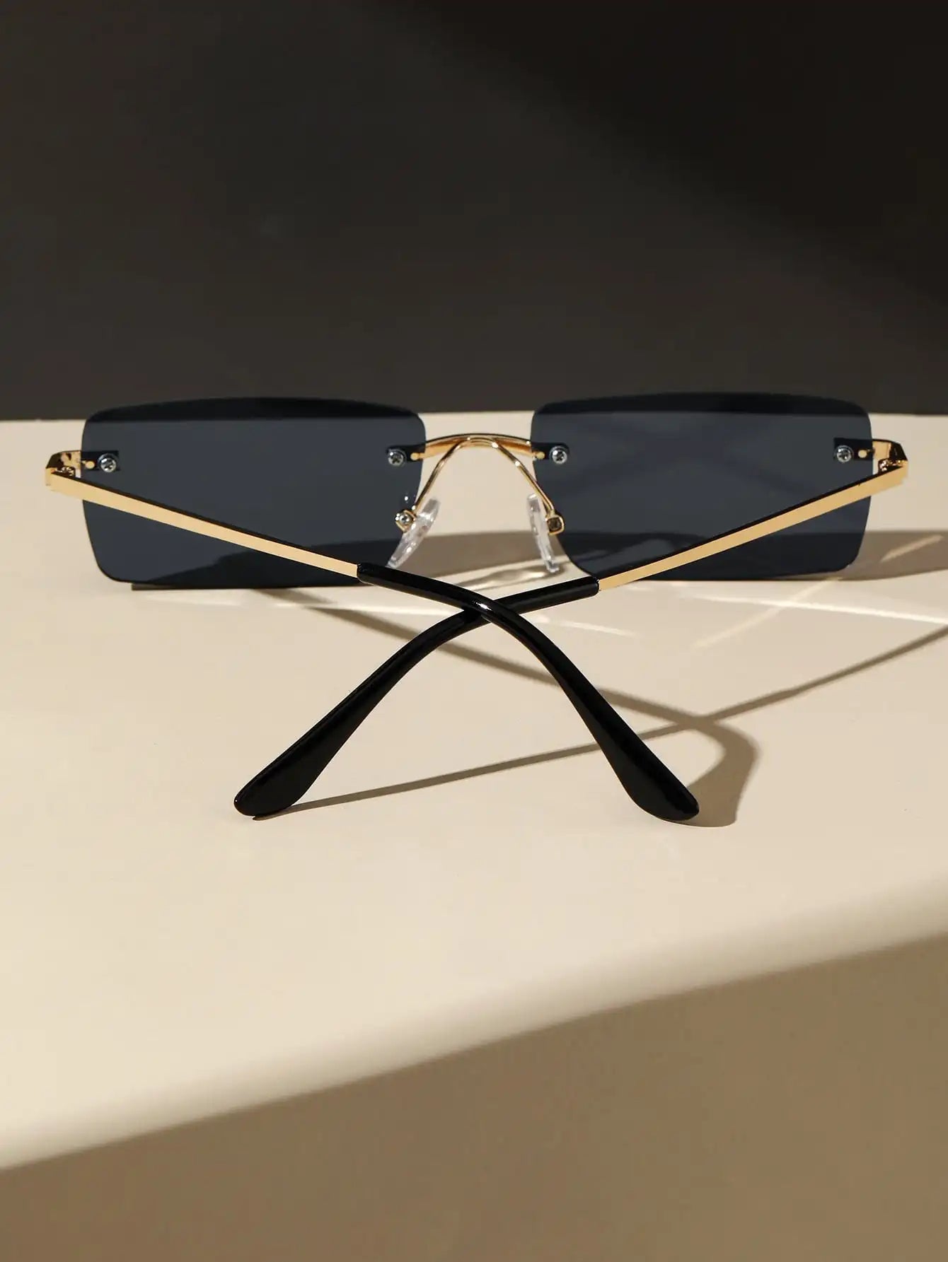 Bondex rimless sunglasses by Sunglass Lover with black lenses and gold arms placed crossed on a beige surface